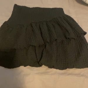 Svart volangkjol från Gina Tricot - Supersöt svart kjol från Gina Tricot med volanger och stretchig midja. Kjolen är kort och har en strukturerad yta som ger extra liv till outfiten. Perfekt att styla med en enkel topp eller stickad tröja.