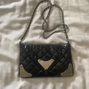 Svart quiltad axelväska med kedjerem - Snygg svart quiltad axelväska med silverfärgade metalldetaljer och kedjerem. Väskan har ett kuvertformat lock och dekorativa metallhörn. Perfekt för att lyfta din outfit med en edgy touch.