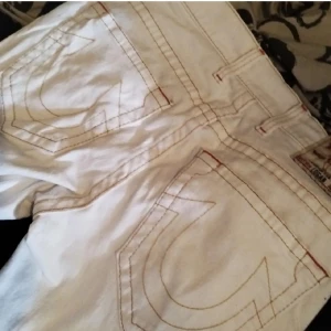Vita jeansbyxor från True Religion - Säljer ett par vita jeansbyxor från True Religion med klassiska stora bakfickor och kontrastsömmar. Byxorna har normal passform och raka ben. Perfekta för dig som vill sticka ut med en ikonisk denimstil.