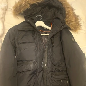 Svart parkajacka med fuskpäls - Svart parkajacka från Hollister med huva och fuskpäls. Jackan har flera fickor framtill, dragkedja och knappar. Perfekt för kalla dagar med sin varma och rejäla design.