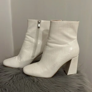Vita boots med krokodilpräglat mönster och klack - Säljer ett par trendiga vita boots med krokodilpräglat mönster och kraftig klack. Skorna har dragkedja på sidan och rund tå. Perfekta för dig som vill sticka ut med en stilren look.