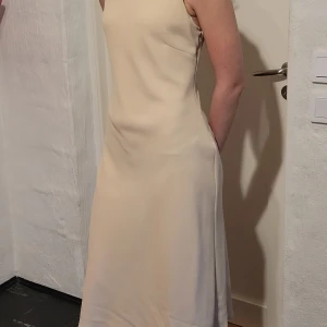 Champagnefärgad långklänning från Randigarutan - Elegant champagnefärgad långklänning från Randigarutan. Klänningen är ärmlös med en stilren, slät design och mjukt fallande material. Perfekt för dig som vill ha en minimalistisk och tidlös look.