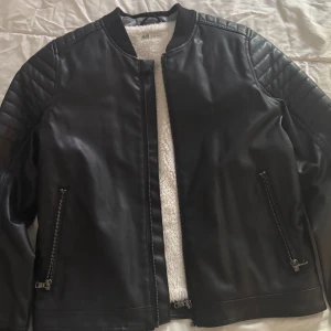 Svart skinnjacka från H&M - Snygg svart skinnjacka från H&M med quiltade detaljer på axlarna och dragkedjor vid fickorna. Jackan har en rak passform och är fodrad med mjukt vitt tyg på insidan. Perfekt för en cool och stilren look. Passar till barn 9-10 år. EUR 140.