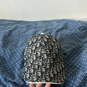 Mönstrad keps från Dior - Säljer en snygg keps från Dior med svart och grått mönster över hela kepsen. Kepsen har böjd skärm och justerbart spänne bak. Perfekt accessoar för att lyfta din outfit.
