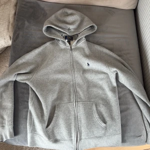 Grå hoodie med dragkedja från Polo Ralph Lauren - Säljer en klassisk grå hoodie från Polo Ralph Lauren med dragkedja och huva. Tröjan har två fickor framtill och en liten broderad logga på bröstet. Perfekt för en avslappnad stil.