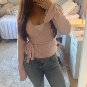 Ljusrosa omlottopp med knytband - Säljer en ljusrosa omlottopp med långa ärmar och knytband i midjan. Toppen har en feminin v-ringning och är i mjukt material, perfekt att styla med jeans eller kjol.