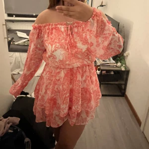 Rosa och vit offshoulder klänning med volang - Säljer en söt offshoulder klänning i rosa och vitt med blommigt mönster. Klänningen har långa ärmar, volangdetaljer och en luftig, kort passform. Perfekt för sommarens alla tillfällen!