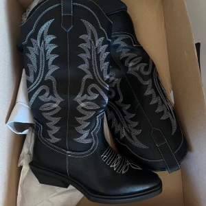 Svarta cowboy boots med vit brodyr - Snygga svarta cowboy boots med vit broderad dekor längs skaftet och foten. Klassisk spetsig tå och låg klack. Perfekta för att lyfta din outfit med western-vibe.