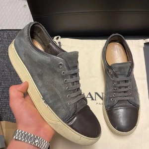 Grå sneakers från Lanvin med mockadetaljer - Säljer ett par grå sneakers från Lanvin med ovandel i mocka och tåparti i mörkare skinn. Skorna har platt, vit sula och klassisk snörning. Perfekta för en stilren och avslappnad look.