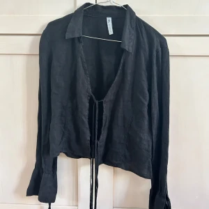 Svart croppad linneskjorta från Zara - Svart croppad skjorta från Zara i linne med långa ärmar och knyt framtill och vid ärmarna. Skjortan har klassisk krage och en avslappnad passform, perfekt för en trendig look.