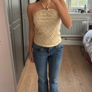 Beige randig halterneck tubtopp s-xs - Säljer en beige tubtopp med tunna diagonala ränder och halterneck med ringdetalj framtill. Toppen knyts i nacken och har en lätt glansig finish. Perfekt för varma dagar och enkel att styla med jeans eller kjol.