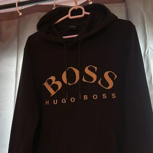 Svart hoodie från Hugo Boss - Säljer en svart hoodie från Hugo Boss med stor guldfärgad logga på bröstet. Tröjan har huva med dragsko och en stor magficka framtill. Perfekt för en stilren och avslappnad look.