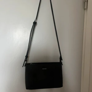 Svart axelväska från Calvin Klein - Säljer en stilren svart axelväska från Calvin Klein med justerbart axelband och dragkedja upptill. Väskan har en minimalistisk design med diskret logga framtill och passar perfekt för att bära det viktigaste.