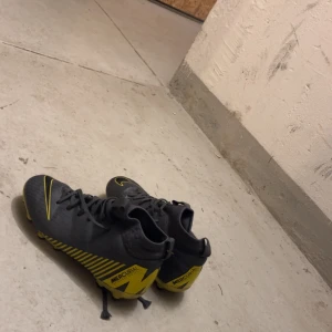 Nike Mercurial fotbollsskor svart/gul - Säljer ett par Nike Mercurial fotbollsskor i svart med gula detaljer och snörning. Skorna har rund tå, platt sula med dobbar och är perfekta för dig som vill sticka ut på planen. Snygg design med stor gul Nike-logga på ovansidan.