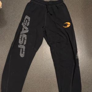 Svarta byxor från Gasp med tryck - Svarta mjukisbyxor från Gasp med elastisk midja och dragsko. Stora gråa tryck på ena benet och ett orange logotryck på höften. Perfekta för träning eller chill. Mjukt material och avslappnad passform.