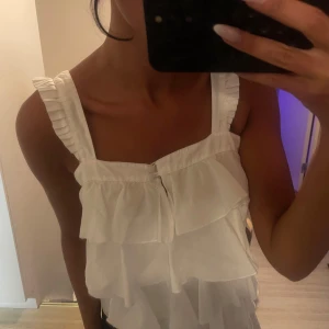 Vit volanglinne med breda axelband - Säljer ett vitt linne med flera lager volanger och breda axelband med rynkade detaljer. Toppen har en söt knappdetalj framtill och är perfekt för varma dagar.