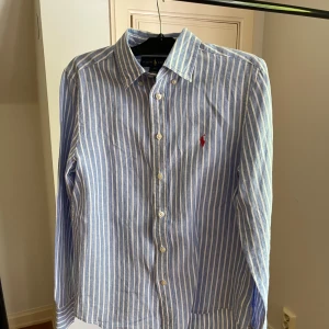 Blåvit randig skjorta från Ralph Lauren - Klassisk blå och vit randig skjorta från Ralph Lauren med broderad röd logga på bröstet. Skjortan har knappar framtill och krage, perfekt för en stilren look.