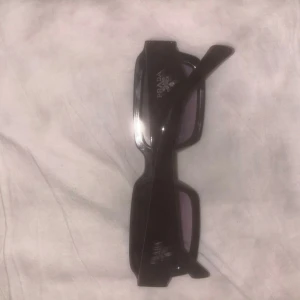 Svarta solglasögon från Prada - Snygga svarta solglasögon från Prada med rektangulära bågar och mörka glas. Klassisk och stilren design med diskret logga på insidan av skalmen. Perfekta för dig som vill ha en tidlös accessoar med lyxig känsla.