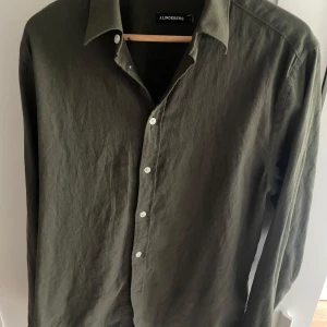 Olivgrön skjorta från J.Lindeberg - Säljer en stilren olivgrön skjorta från J.Lindeberg. Skjortan har klassisk krage, långa ärmar och knäppning framtill med vita knappar. Perfekt för dig som gillar en enkel och tidlös look.