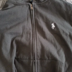 Svart hoodie från Polo Ralph Lauren - Svart hoodie från Polo Ralph Lauren med dragkedja framtill och klassisk vit broderad logga på bröstet. Modellen har huva och fickor på magen. Perfekt för en avslappnad och stilren look.