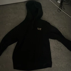 Svart hoodie från Emporio Armani EA7 - Svart hoodie från Emporio Armani EA7 med gul logotyp på bröstet. Klassisk huva med dragsko och mjukt material. Perfekt för en stilren och avslappnad look.