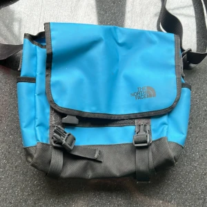 The North Face väska blå - Säljer en blå axelväska från The North Face med svarta detaljer. Väskan har justerbar axelrem, spännen framtill och ett nätfack med dragkedja på insidan. Perfekt för att bära med sig det viktigaste till vardags.