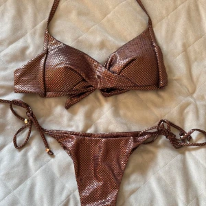 Brun glittrig bikini med knyt - Säljer en snygg brun bikini med glittrigt material och knyt i både överdel och underdel. Bikinin har trekantsformad överdel och smala band med pärldetaljer på sidorna. Perfekt för sommaren! Från Calzedonia, underdelen är i storlek S och överdelen i storlek 80A (tror jag).
