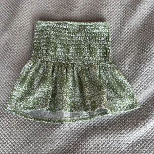 Grön blommig tubtopp från Gina Tricot - Supersöt grön kortkjol från Gina Tricot med vitt blommönster och smock i midjan. Kjolen har en luftig och lätt känsla, perfekt för varma dagar. Passar dig som gillar en feminin och somrig stil!