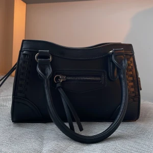 Svart handväska med flätade detaljer - Säljer en svart handväska med snygga flätade detaljer på sidorna och en liten dragkedjeficka framtill. Väskan har både handtag och en avtagbar axelrem för olika stilar. Perfekt storlek för det viktigaste och stilren design. Använt 1 gång då jag fick den i present.  Är från Gina Tricot "crossbody bag". Är hel och fin och som ny. 