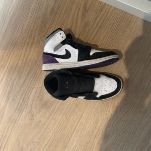 Nike Air Jordan 1 Retro High Court Purple - Säljer ett par Nike Air Jordan 1 Retro High Court Purple med svart, vitt och lila färgblock. Skorna har klassisk snörning, svart swoosh och rund tå. Ovandelen är i skinn och mocka, med lila detaljer på hälen och sulan.