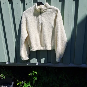 Vit fleece-tröja med halv dragkedja - Mysig vit fleece-tröja med halv dragkedja och hög krage. Tröjan har lång ärm och resår i midjan samt vid ärmsluten för en skön passform. Perfekt för kyliga dagar.