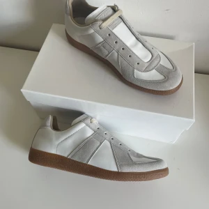 Maison Margiela gats - Säljer ett par vita sneakers från Maison Margiela med beige mockadetaljer och bruna gummisulor. Boxen och dustbagen tillkommer i köpet! Digitalt kvitto finns. Frågor kring skona-> Privat