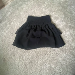 Svart volangkjol med smock i midjan - Supersöt svart kjol med smockad midja och dubbla volanger. Kjolen är kort och har en luftig känsla, perfekt för varma dagar. Passar till många olika stilar och är lätt att matcha med olika toppar. Skriv för fler bilder 🥰