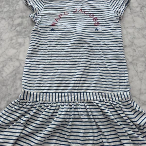 Randig klänning från Little Marc Jacobs - Säljer en söt vit och blå randig klänning från Little Marc Jacobs med röd logotyp framtill. Klänningen har korta ärmar, rund halsringning och en dekorativ dragkedja baktill. Perfekt för en avslappnad och stilren look. Liten fläck se sista bilden går nog att få bort vi har inte försökt! 