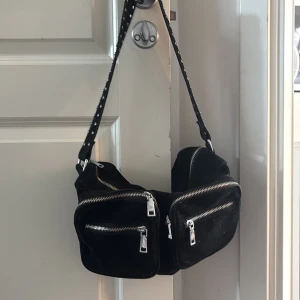 Svart axelväska med nitar och dragkedjor - Cool svart axelväska med bred axelrem dekorerad med silvriga nitar. Väskan har flera fack framtill med silvriga dragkedjor och är tillverkad i ett mjukt material. Kommer även med ett annat band.