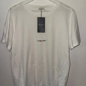Vit t-shirt från Saint Laurent - Säljer en stilren vit t-shirt från Saint Laurent med liten broderad logga på bröstet. T-shirten har rund halsringning och korta ärmar. Perfekt för en clean och klassisk look.