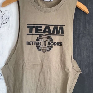 Beige linne från Better Bodies - Säljer ett beige linne från Better Bodies med svart TEAM-tryck och grafiskt motiv framtill. Modellen är ärmlös med djupa ärmhål, perfekt för gymmet eller träning.