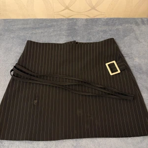 Svart randig kjol med bälte från MissDenis - Säljer en brun kortkjol från MissDenis med tunna ljusa ränder och ett snyggt bälte med silverfärgat spänne. Kjolen har omlottkänsla och är perfekt för dig som gillar stilrena detaljer.