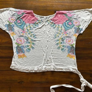 Vit blus med blommigt mönster och rosa spets - Säljer en vit blus med färgglatt blommigt mönster på sidorna och rosa spetsdetaljer vid axlarna. Blusen har korta ärmar och knytband nertill för en extra detalj. Perfekt för dig som gillar bohemisk stil!
