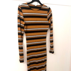 Randig ribbad klänning - Säljer en tight ribbad klänning med svarta, bruna, vita och beigea horisontella ränder. Klänningen har lång ärm och rund halsringning. Perfekt för dig som gillar en figurnära passform och stilrena ränder. Från Nelly. 