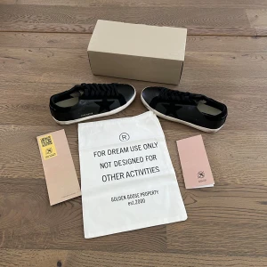 Svarta sneakers från Golden Goose - Säljer ett par svarta sneakers från Golden Goose med vit sula och svart stjärndetalj på sidan. Skorna har snörning och klassisk rund tå. Märkeslogga syns baktill. Perfekta för dig som gillar stilrena och trendiga sneakers.