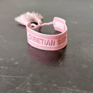 Rosa armband från Christian Dior - Säljer ett ljusrosa armband från Christian Dior med broderad logga och två tofsar. Armbandet är justerbart och har en mjuk textilstruktur som ger en trendig och avslappnad look.