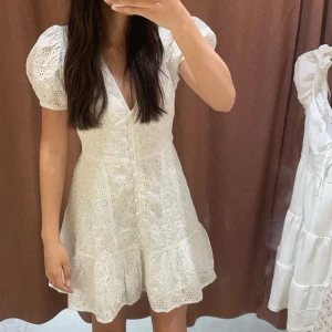 Vit kortklänning med puffärm och broderade detaljer - Superfin vit kortklänning med v-ringning 🤍, puffärmar och broderade blommor. Klänningen har knappar framtill och volang nertill. Perfekt till sommaren!