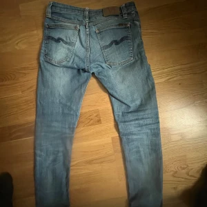 Blå jeansbyxor från Nudie Jeans - Säljer ett par klassiska blå jeans från Nudie Jeans med tydliga slitningar och Nudie-loggan på bakfickorna. Jeansen har normal passform och raka ben, samt detaljer som nitar och orange sömmar.