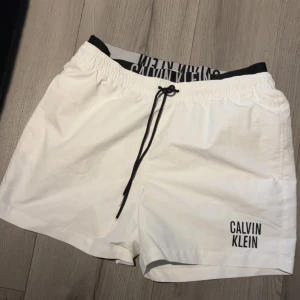 Vita badshorts från Calvin Klein - Snygga vita badshorts från Calvin Klein med svart snörning i midjan och logga tryckt på ena benet. Shortsen har elastisk midja och innerbyxa i mesh för extra komfort. Perfekta för stranden eller poolen.