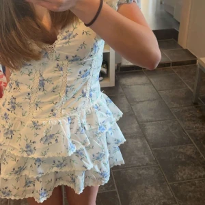 Vit blommig volangklänning - Supersöt kortklänning i vitt med blått blommönster och volanger. Klänningen har v-ringning, spetsdetaljer och puffiga kortärmar. Perfekt för sommarens alla tillfällen!