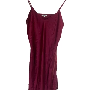 Vinröd satin klänning från Change - Vinröd långklänning i satin från Change med smala axelband och v-ringning. Klänningen har en elegant och enkel design som ger en lyxig känsla. Perfekt för dig som vill ha en stilren look med lite extra glans.