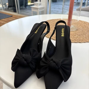 Svarta pumps med rosett och spetsig tå - Säljer ett par helt nya eleganta svarta pumps med spetsig tå och stor dekorativ rosett framtill. Skorna har öppen häl och smal klack, perfekta för att lyfta din outfit.