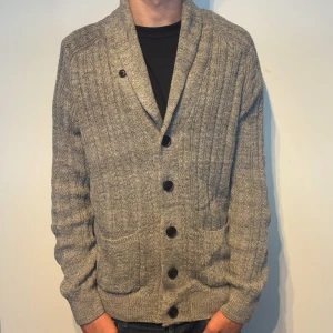 Stickad Cardigan med Fickor - Säljer en grå stickad kofta med kabelstickat mönster, stora knappar framtill och två fickor. Koftan har bred krage och bruna elbow-patches. Perfekt för lager-på-lager och snygg till jeans. Har ni några frågor eller funderingar? - Hör av er!