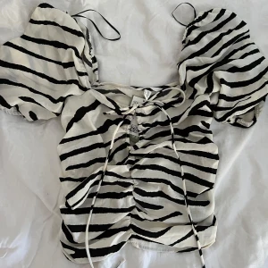 Zebramönstrad topp med puffärmar och knyt - Säljer en vit och svart zebramönstrad topp med stora puffärmar och knyt framtill. Toppen har en croppad passform och rynkade detaljer längs mitten. Perfekt för dig som vill sticka ut med ett trendigt djurmönster.